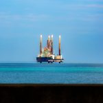 Plataforma de petróleo vista à distância em meio ao oceano, sob céu azul.