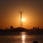 Torre de transmissão elétrica em silhueta ao pôr do sol, diante do reflexo de uma cidade na água.