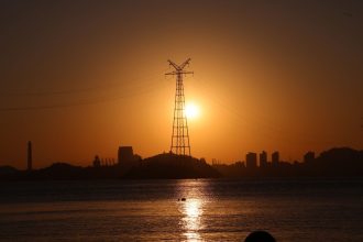 Torre de transmissão elétrica em silhueta ao pôr do sol, diante do reflexo de uma cidade na água.